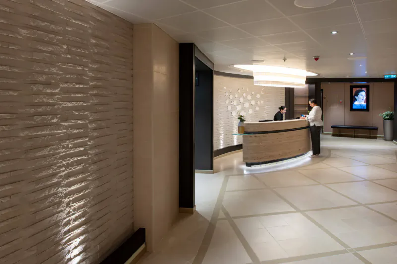 Modern cruise ship reception desk with two uniformed staff welcoming guests, elegant beige interior and plants.