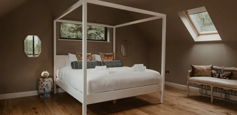 Elegant white four-poster bed with pillows and towels in cozy loft bedroom with skylights and lake view windows, The Boathouse Wales.