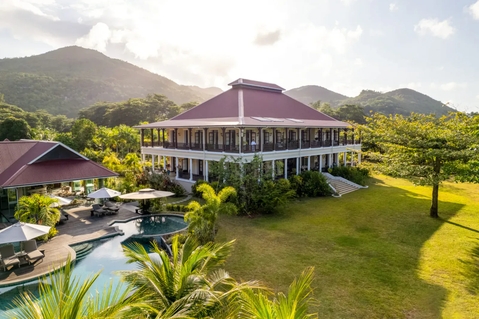 La Cigale Estate on Praslin, Seychelles: red-roofed white colonial house, infinity pool, lounge areas, palms, lush green lawn, mountains at sunset.