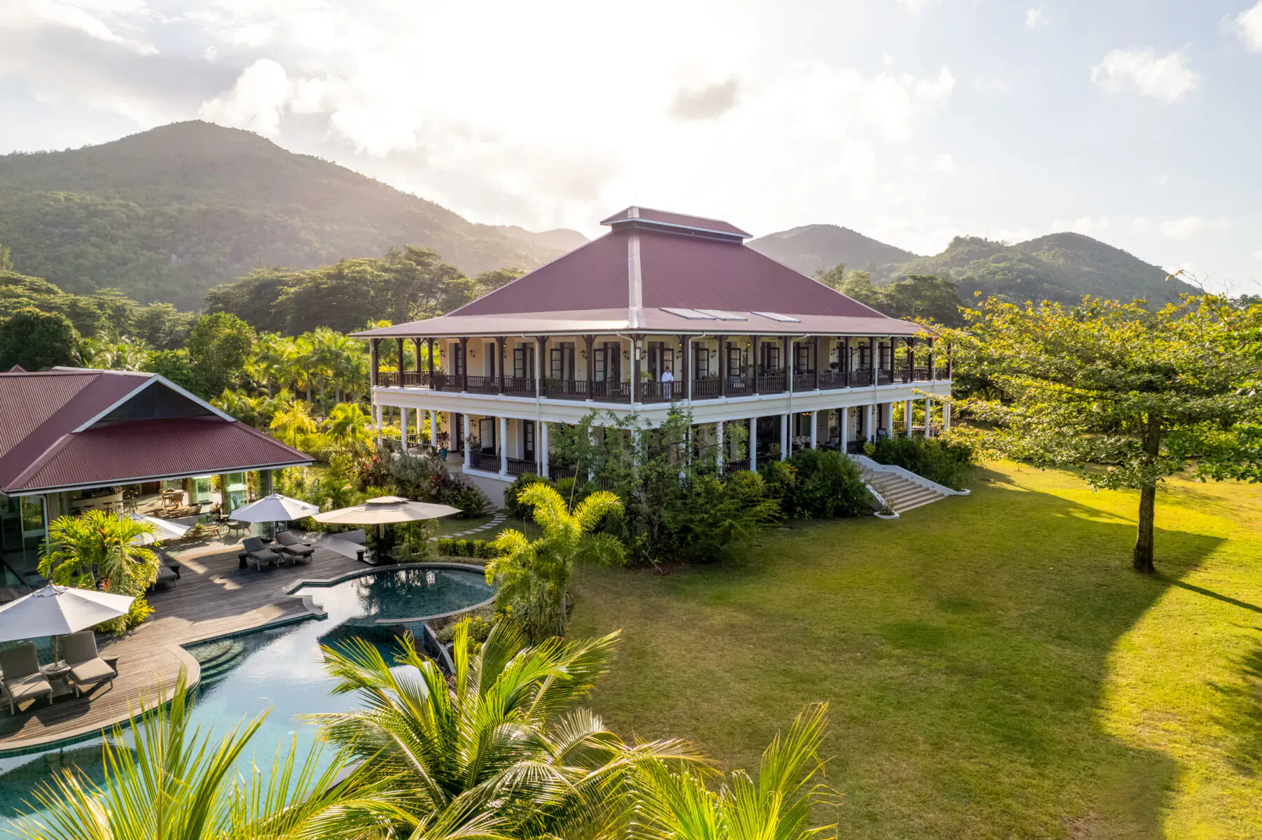 La Cigale Estate on Praslin, Seychelles: red-roofed white colonial house, infinity pool, lounge areas, palms, lush green lawn, mountains at sunset.