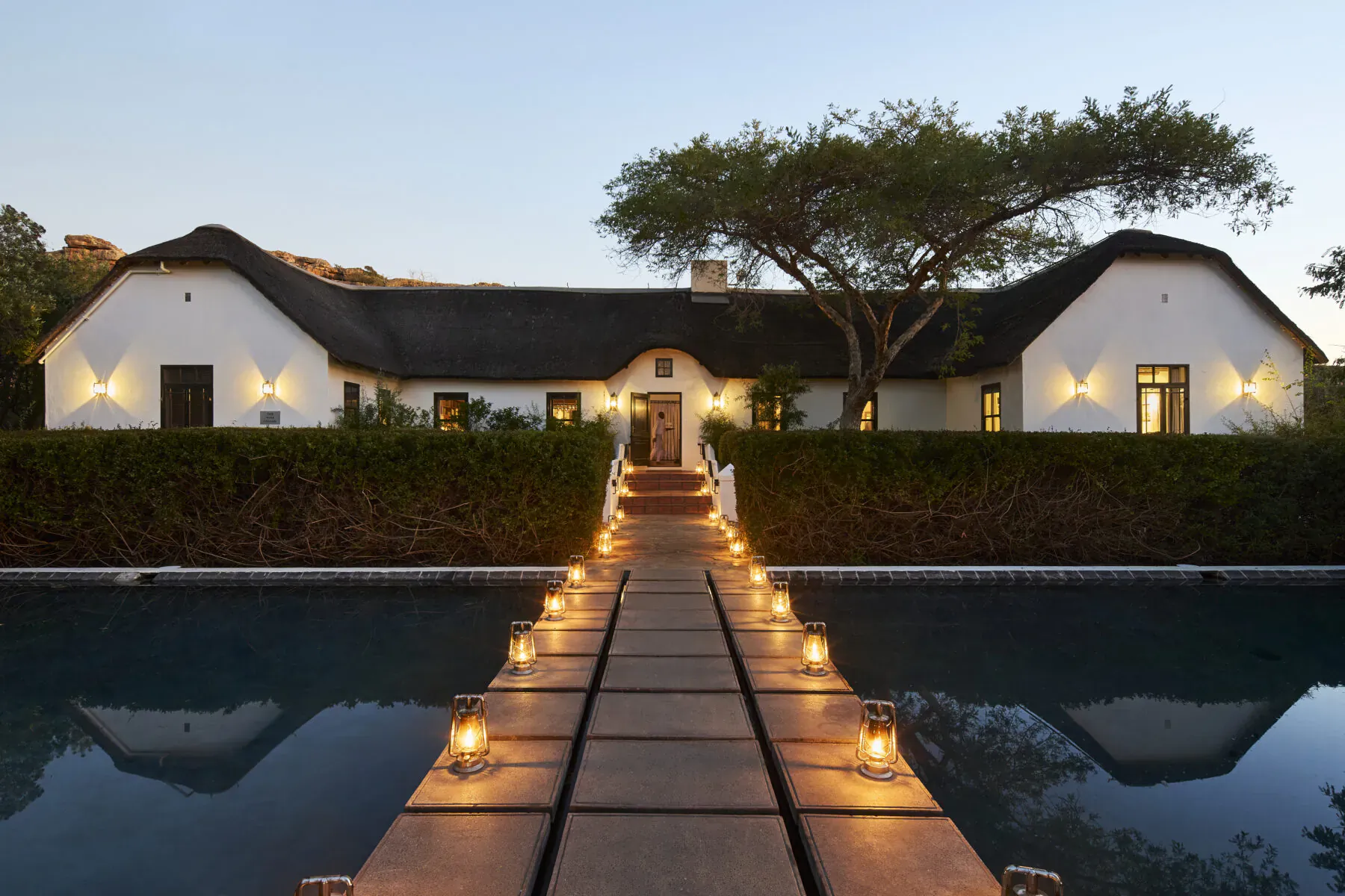 Thatched white luxury lodge at Bushmans Kloof, lit pathway of candles over reflecting pool at twilight under acacia trees.