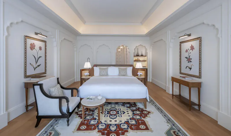 Luxurious hotel bedroom with king bed, ornate rug, armchairs, and floral artwork in elegant white room.
