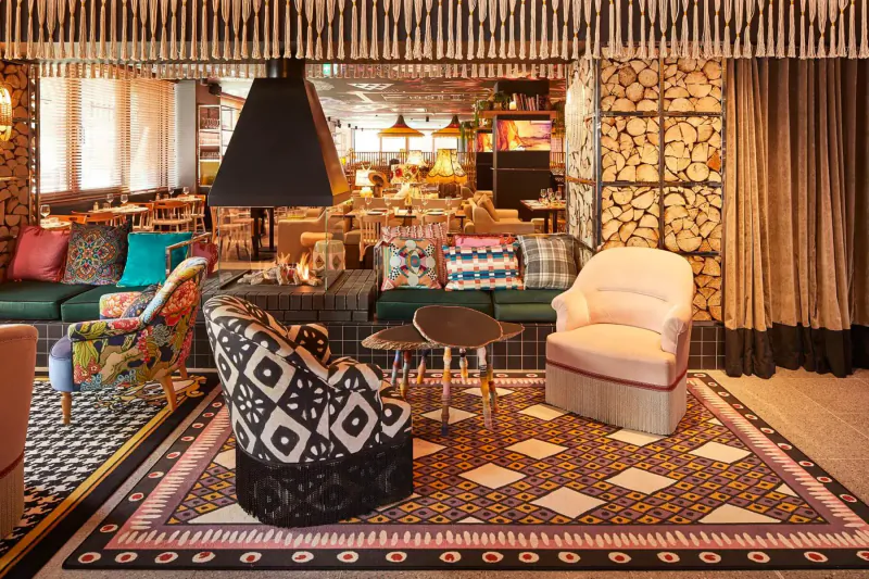 Eclectic hotel lounge with colorful patterned armchairs, sofas, wood walls, lamps, and patterned rug.