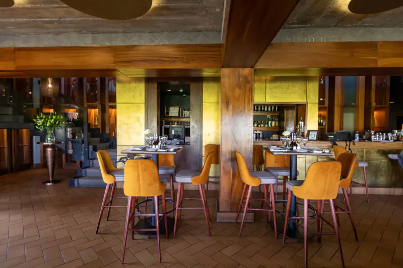 Modern Polo Club restaurant interior with orange upholstered bar stools, wooden beams, plants, and set tables.
