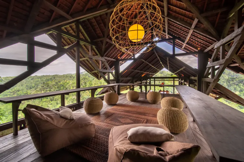 Spacious open-air balcony at Buahan Bali wellness retreat with woven hanging lamp, cushioned daybed, rattan balls, overlooking lush green valley.