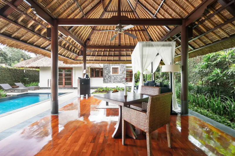 Luxurious open-air Balinese pavilion with thatched roof, wooden dining table, pool, and tropical forest views in Ubud.