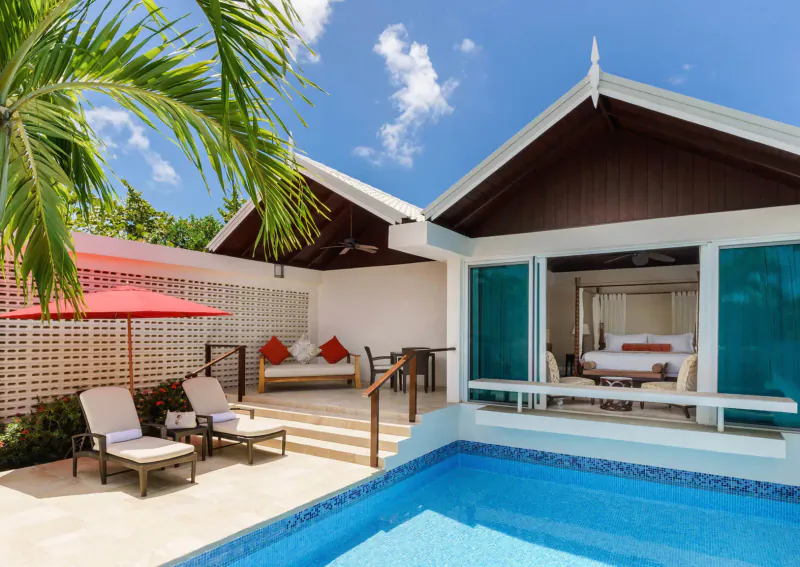 Luxury beachfront villa at Spice Island Beach Resort, Grenada, with infinity pool, lounge chairs, red umbrella, and bedroom visible through open doors amid palms.