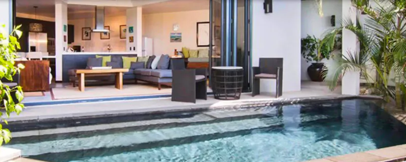 Luxury outdoor patio at Tamarind Hills Antigua with infinity pool, gray sofa, chairs, and open kitchen beyond sliding doors