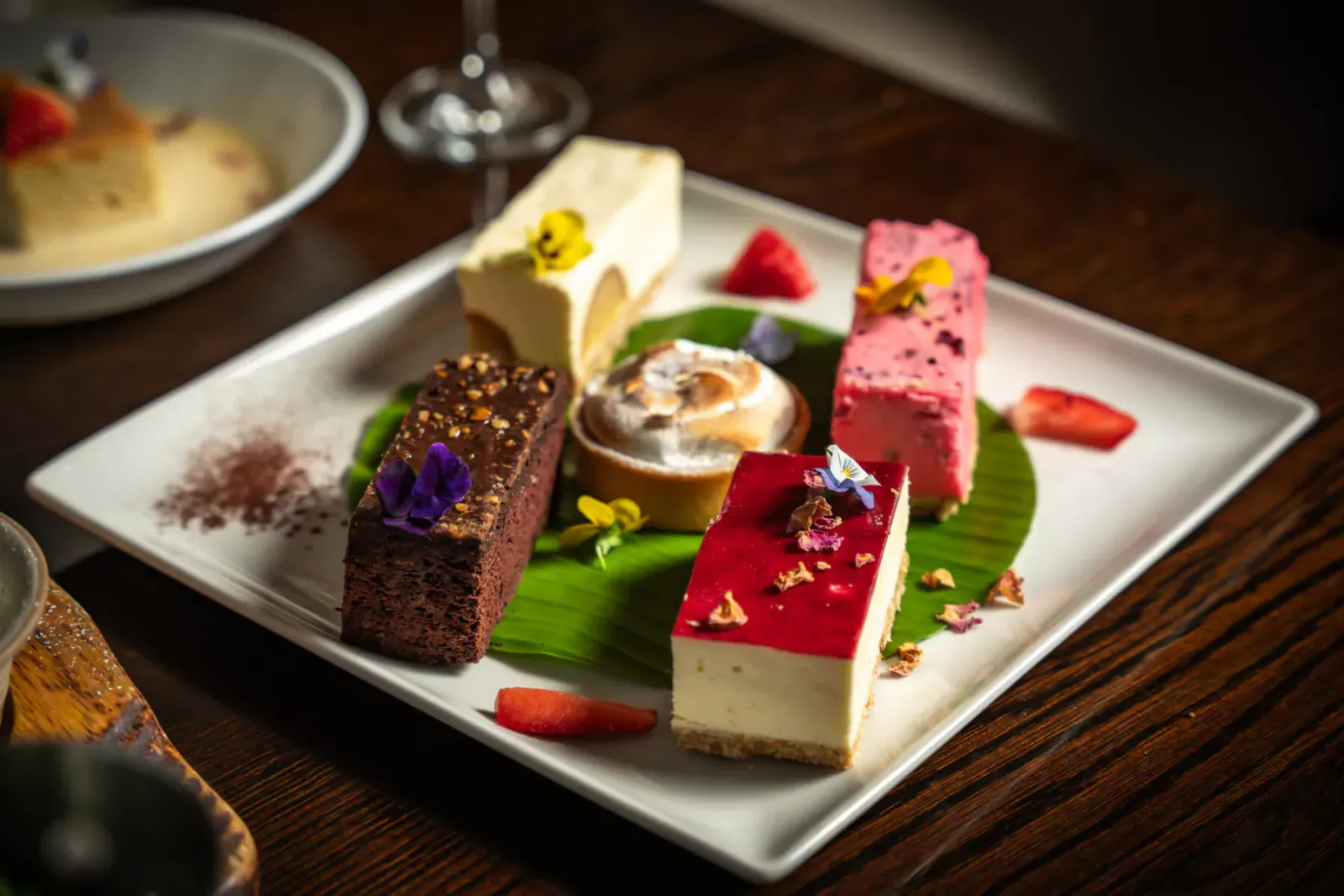 Assortment of gourmet desserts on square plate with edible flowers and betel leaves, beside wine glass at Mint Leaf London.