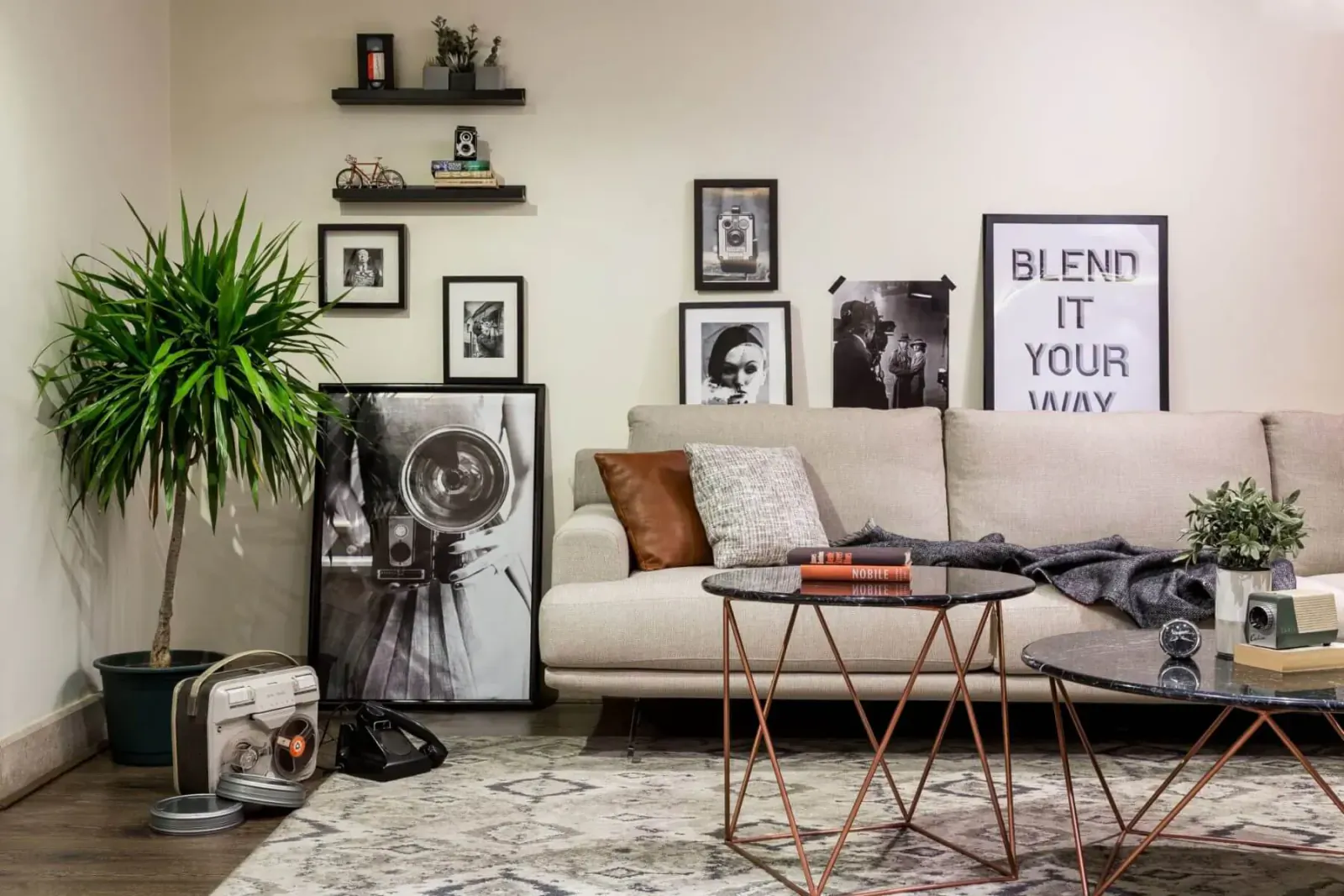 Modern living room with beige sofa, brass coffee table, plants, black-and-white art including 'BLENDO IT YOUR WAY' poster, and vintage suitcase.