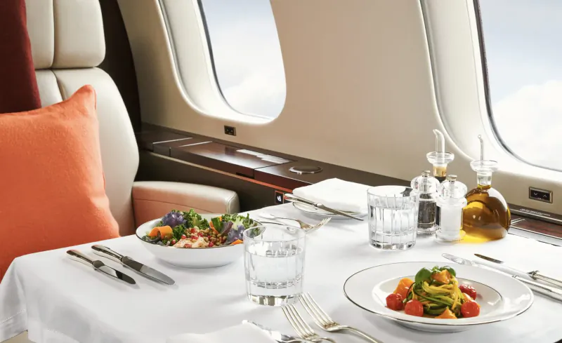 Luxury private jet dining table with fresh vegetable salads, water glasses, oil, and cloudy sky view through windows.