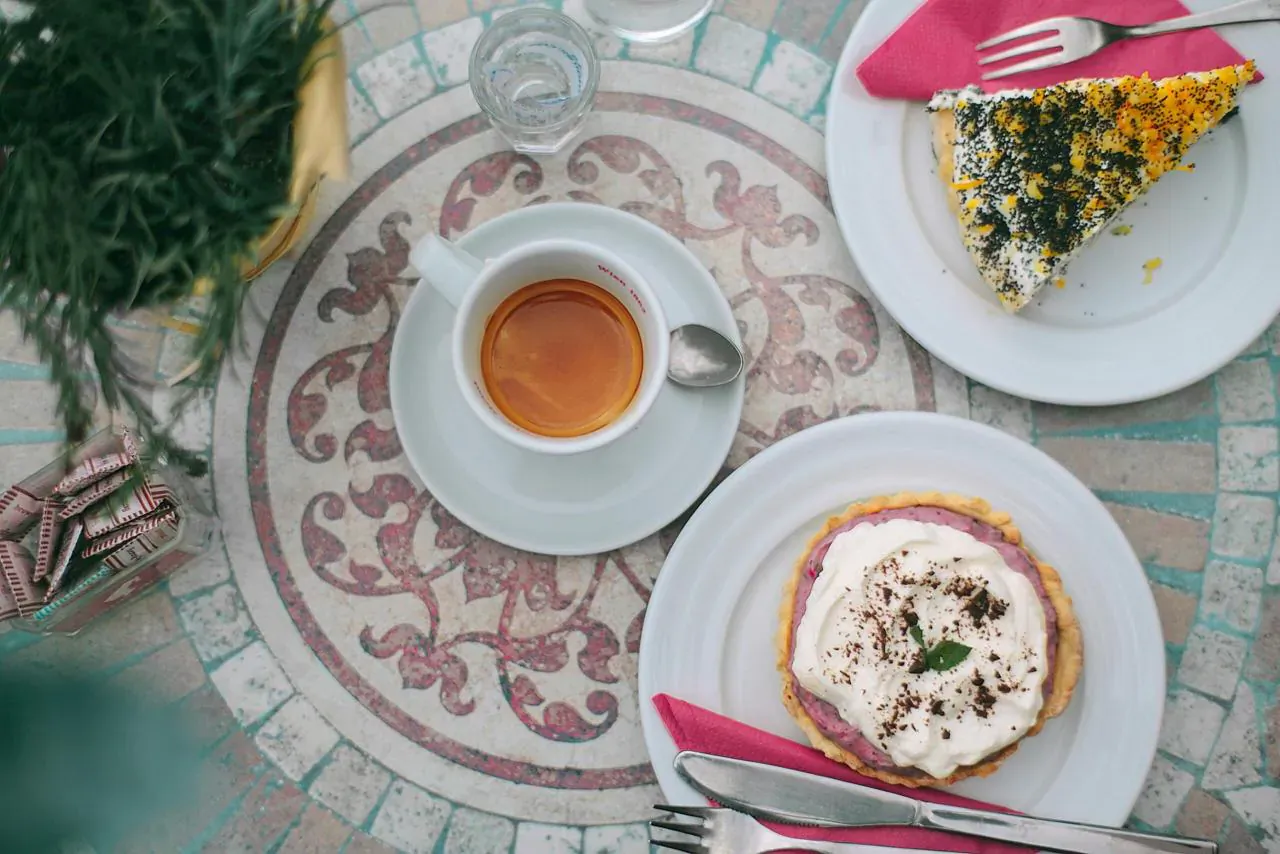 Overhead view of gourmet tea with spoon, whipped cream dessert with mint, and black sesame cake on ornate tiled table.