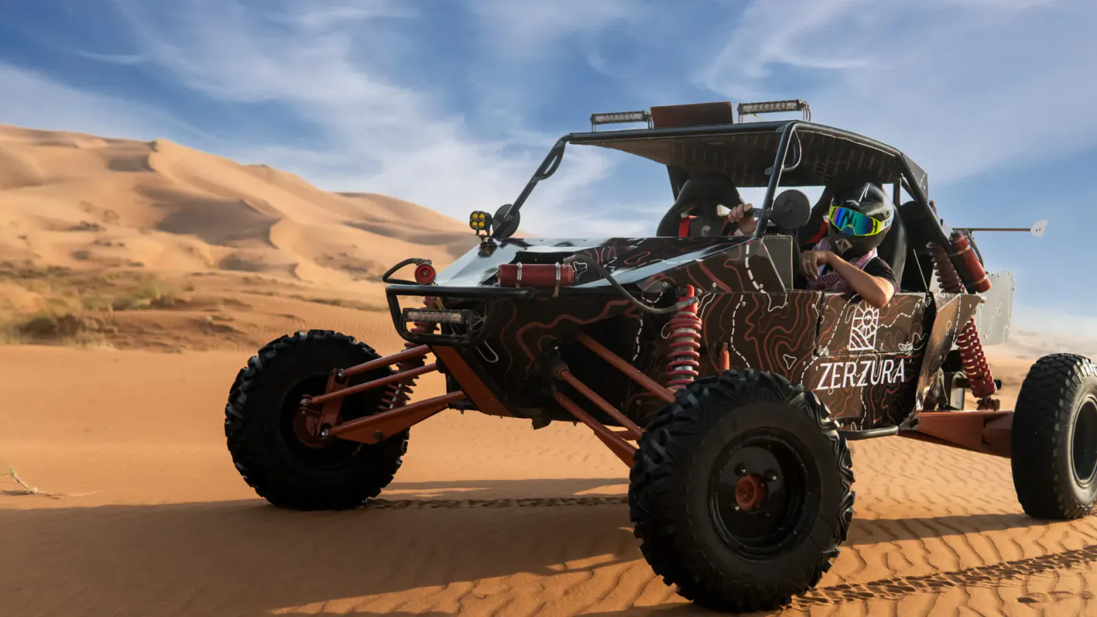 Red Zerzura dune buggy with helmeted drivers speeding through sandy desert dunes under blue sky