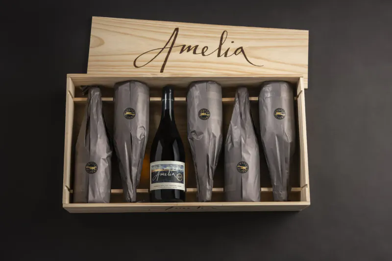 Open wooden box labeled 'Ameliaia' holding six wrapped wine bottles, one partially unwrapped showing 'Amelia' label.