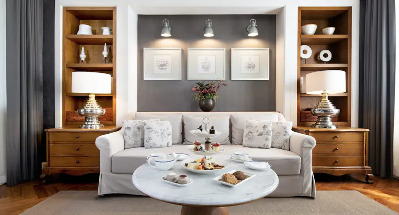 Elegant living room at The Sanchaya with white sofa, round table of pastries, flanked by wooden shelves, lamps, and art on gray wall.
