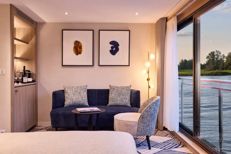 Luxurious cruise ship cabin with navy sofa, armchair, side table, minibar, abstract art, and panoramic river sunset view.