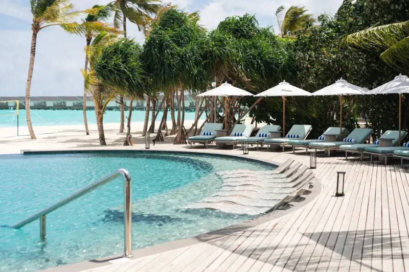Curved turquoise pool with white loungers under palm trees and umbrellas at Raaya by Atmosphere Maldives beach resort