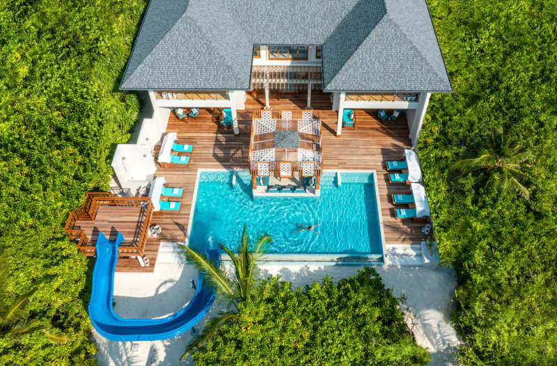 Aerial view of Hideaway Beach Resort villa with infinity pool, blue waterslide, wooden decks, and lounge chairs amid lush greenery.
