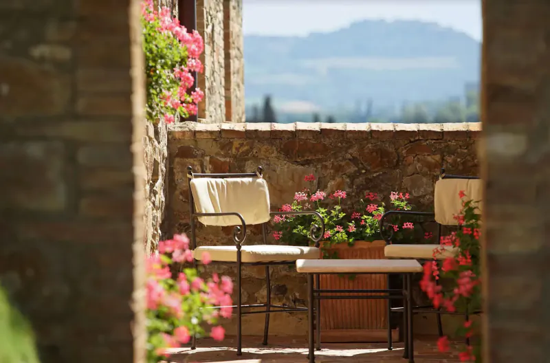 Tuscan stone terrace with two beige chairs, pink flower pots, and distant vineyard hills.