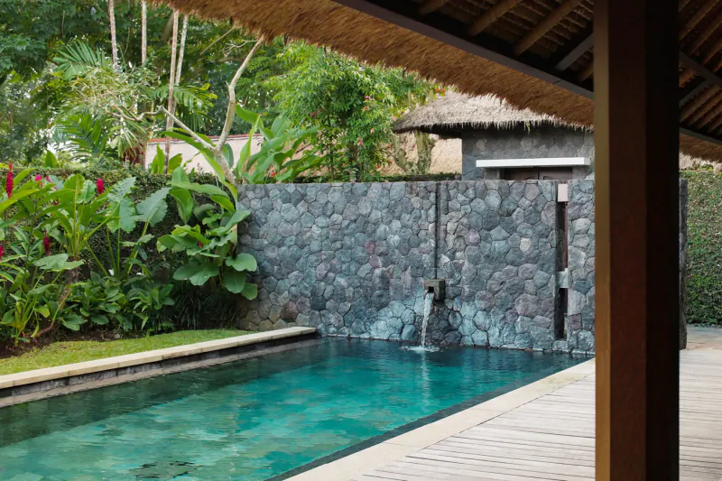 Tropical Bali villa infinity pool with stone waterfall, lush greenery, thatched pavilion overlooking forest.