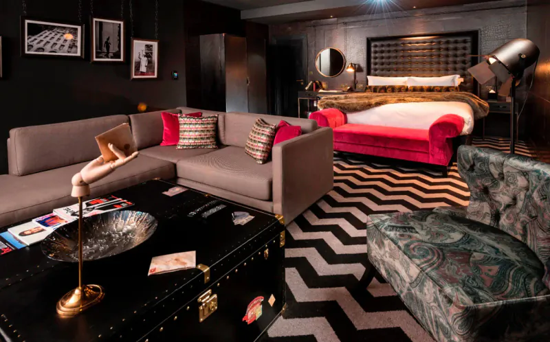 Luxurious Hotel Gotham room with gray L-shaped sofa, pink velvet bed, black zig-zag rug, brass lamps, and vintage trunk coffee table.
