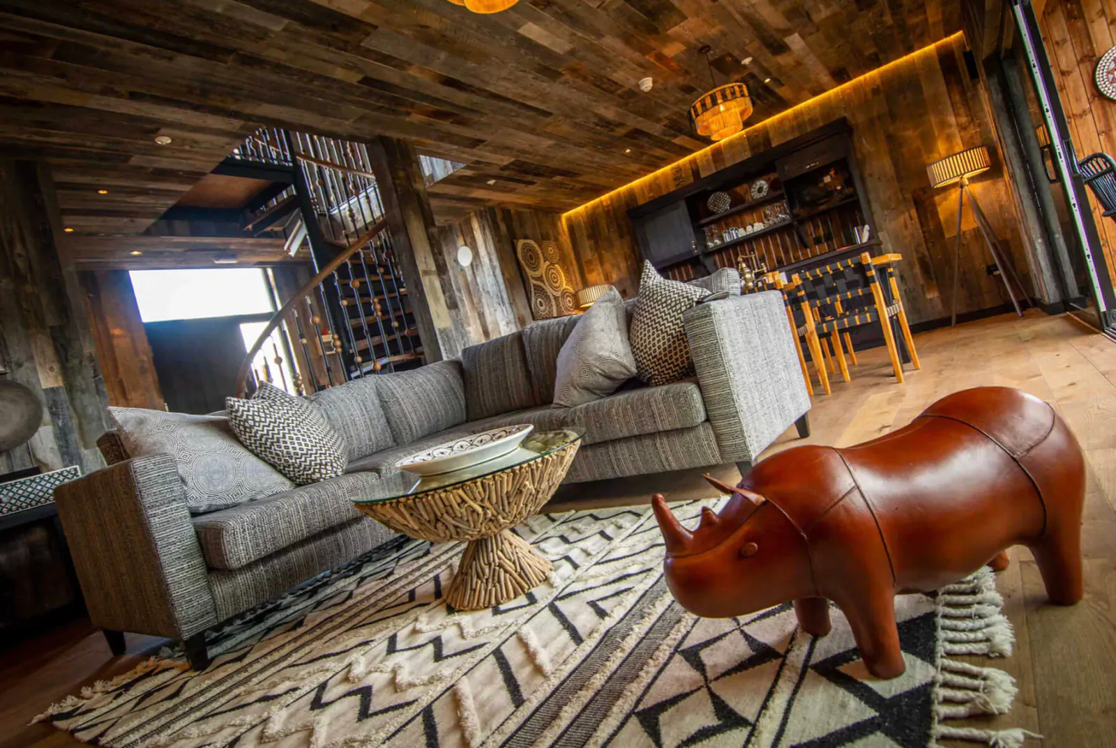 Cozy Rhino Lodge living room with gray sofa, rhino stool on zigzag rug, wood beam ceiling, African decor.