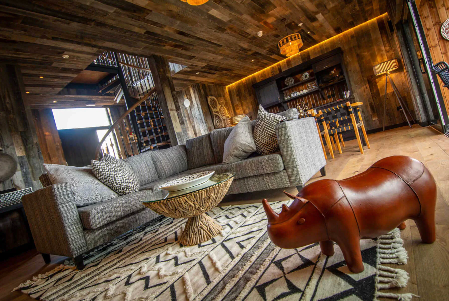 Cozy Rhino Lodge living room with gray sofa, rhino stool on zigzag rug, wood beam ceiling, African decor.