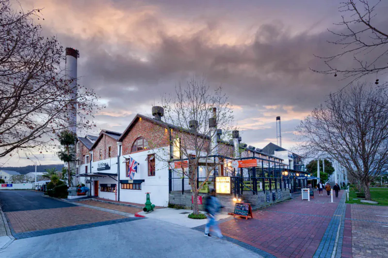 Turbine Boutique Hotel and Spa on Thesen Islands, Knysna, at sunset with brick warehouses, smokestacks, and people walking nearby.