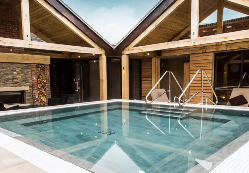 Indoor spa pool with turquoise water, wooden beams, stone fireplaces, and lounge chairs at Moddershall Oaks.