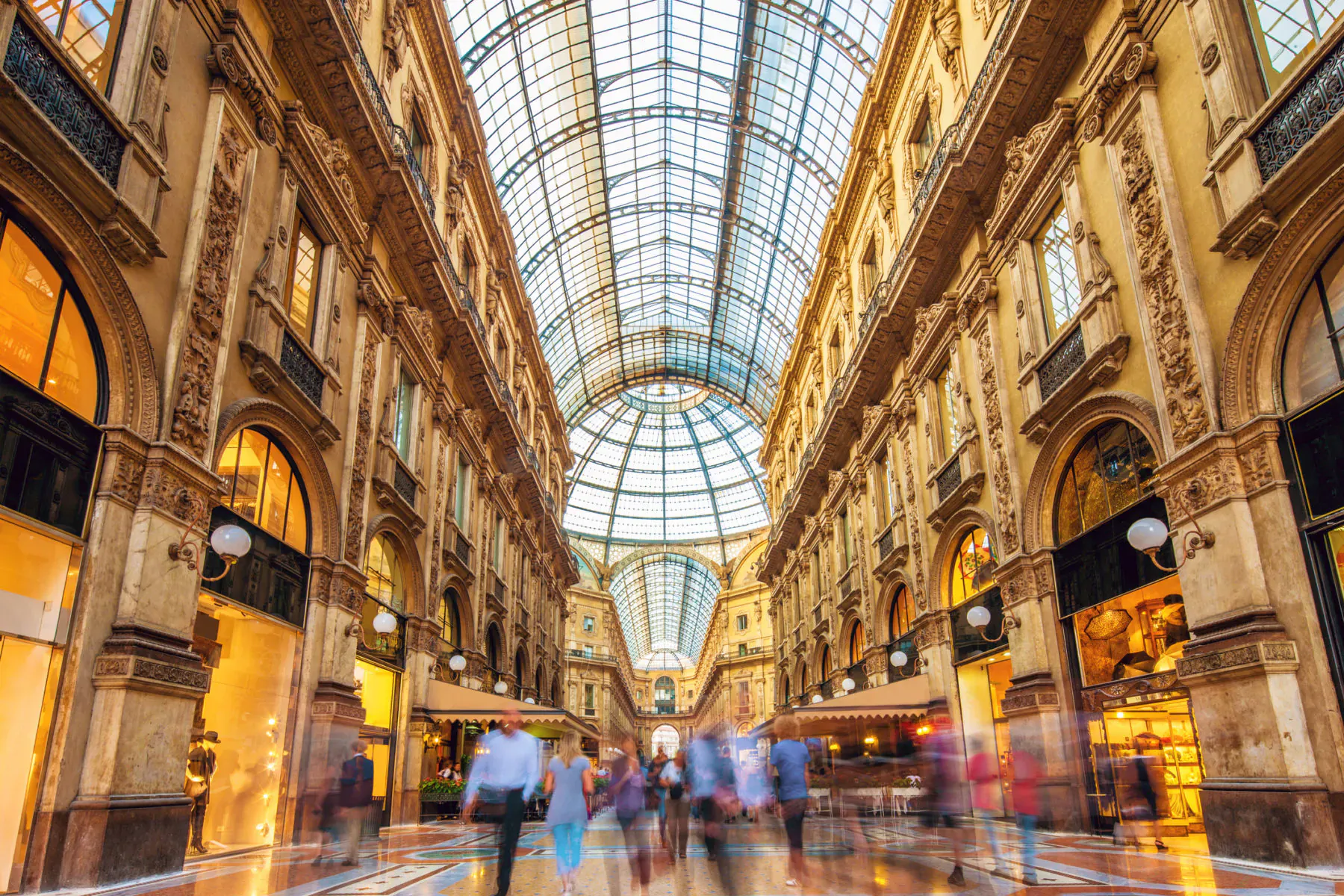 Galleria Vittorio Emanuele II: grand arcade with glass dome, ornate arches, luxury shops, blurred pedestrians walking.