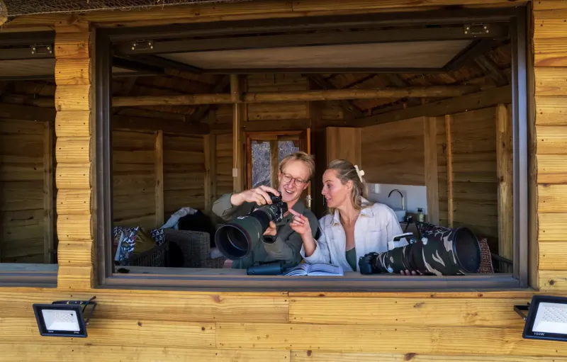 Two women in wooden cabin window, one photographing with camera and tripod, wildlife gear nearby, wilderness setting.