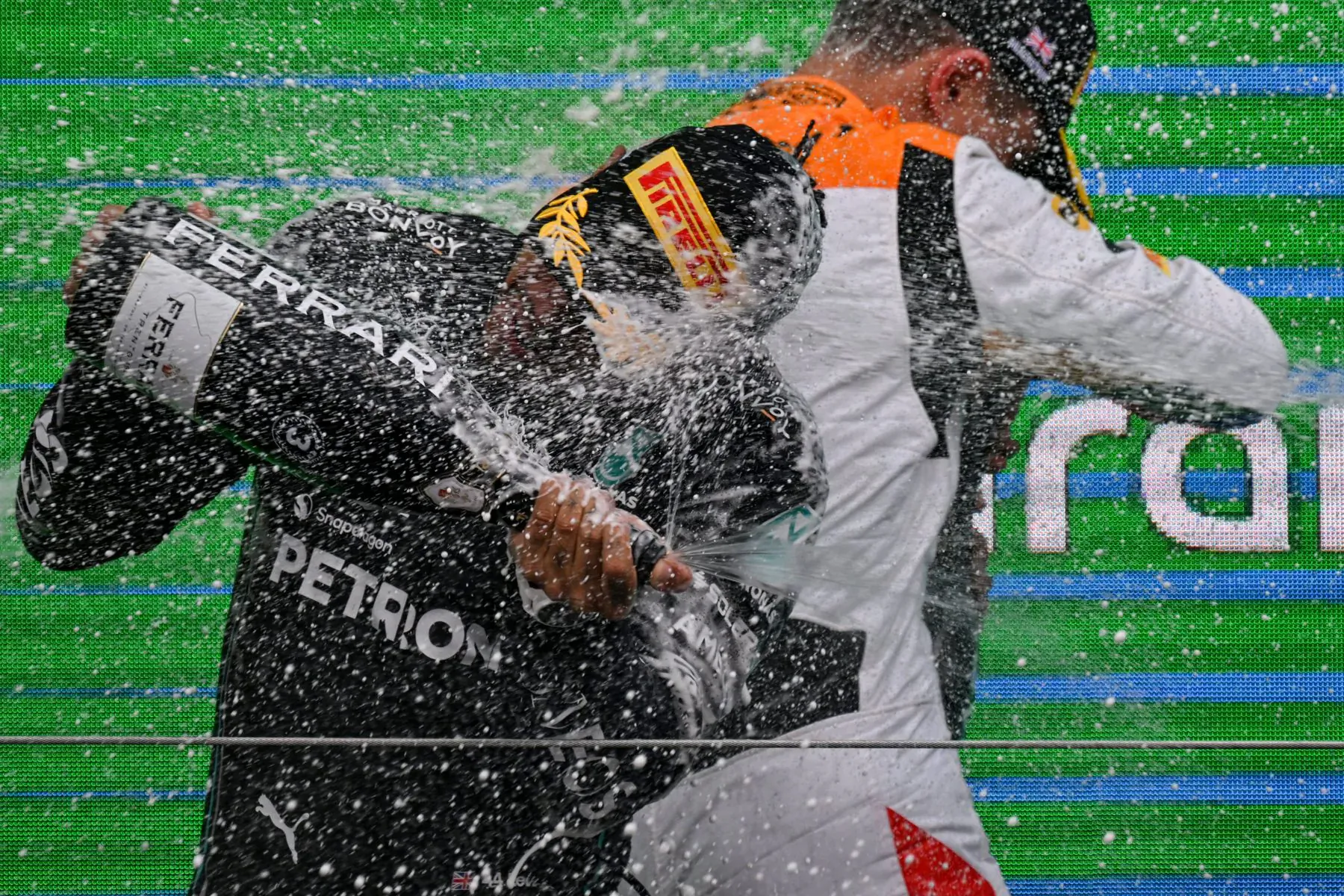 F1 drivers in Mercedes and McLaren suits sprayed with Ferrari Trento champagne on podium amid green backdrop