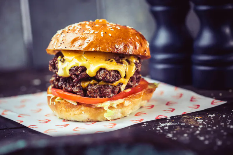 Double cheeseburger with beef patties, melted cheese, lettuce, tomato on sesame bun, on red paper with salt and pepper shakers