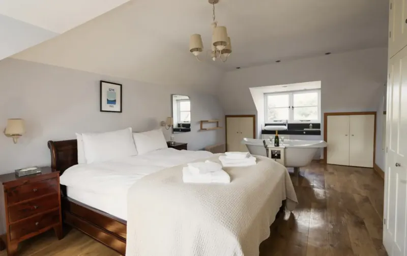 Spacious attic bedroom in Cotswolds country house with king bed, freestanding bathtub, champagne bottle, and skylights.