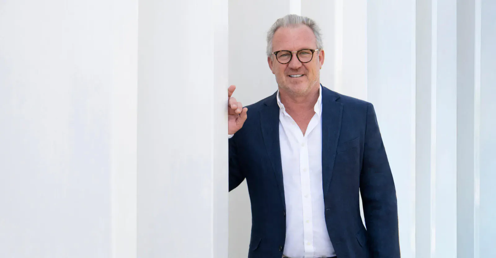 Smiling man with glasses in navy suit and white shirt, peeking from behind white pillar