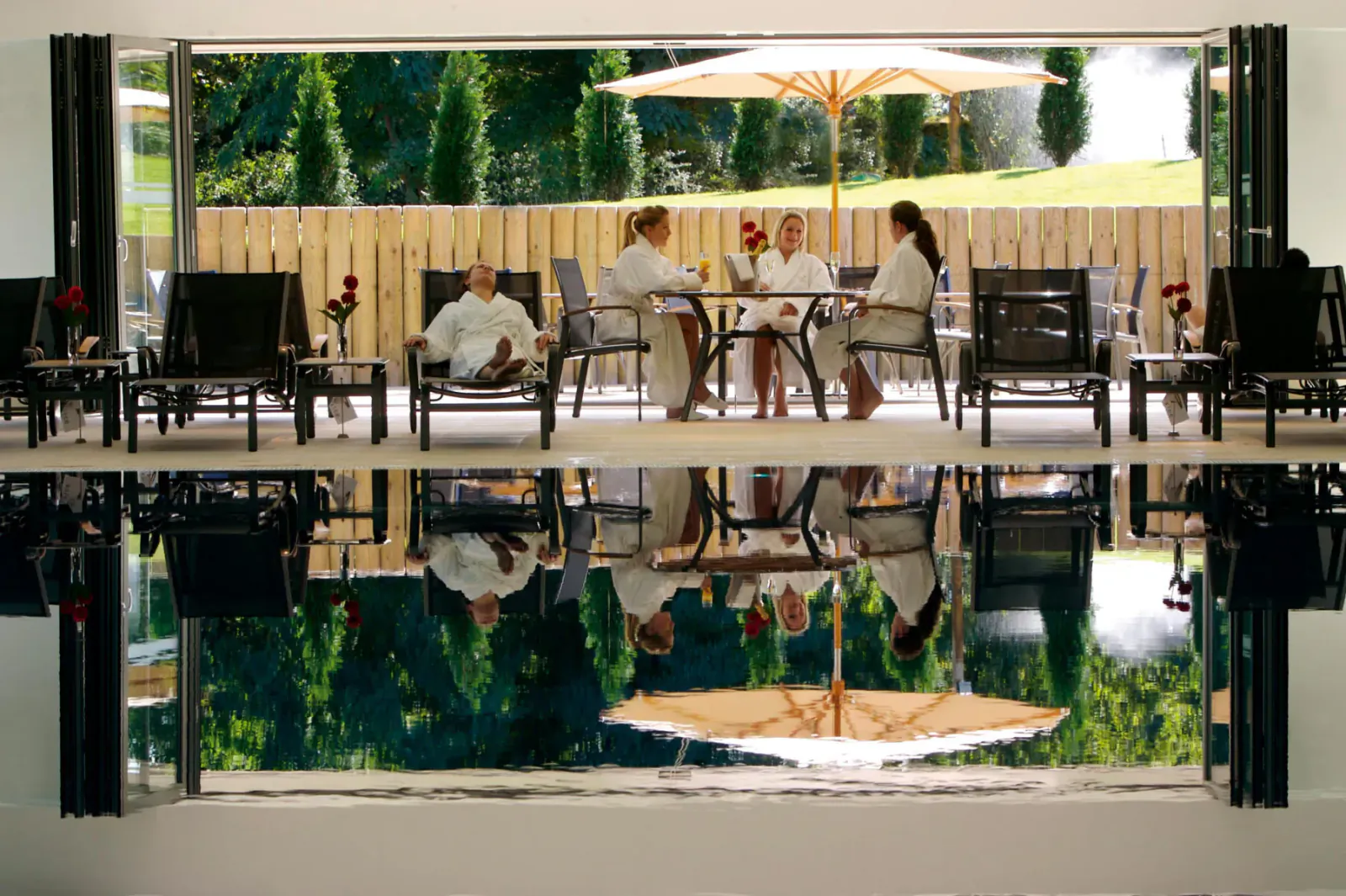 Four people in white robes relax at a table on a terrace by infinity pool reflecting lush green garden and umbrella, Donnington Valley.