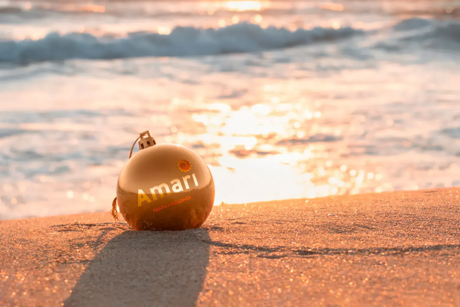 Golden 'Amari' ornament on sandy beach with ocean waves at sunset, for Amari Raaya Maldives NYE event