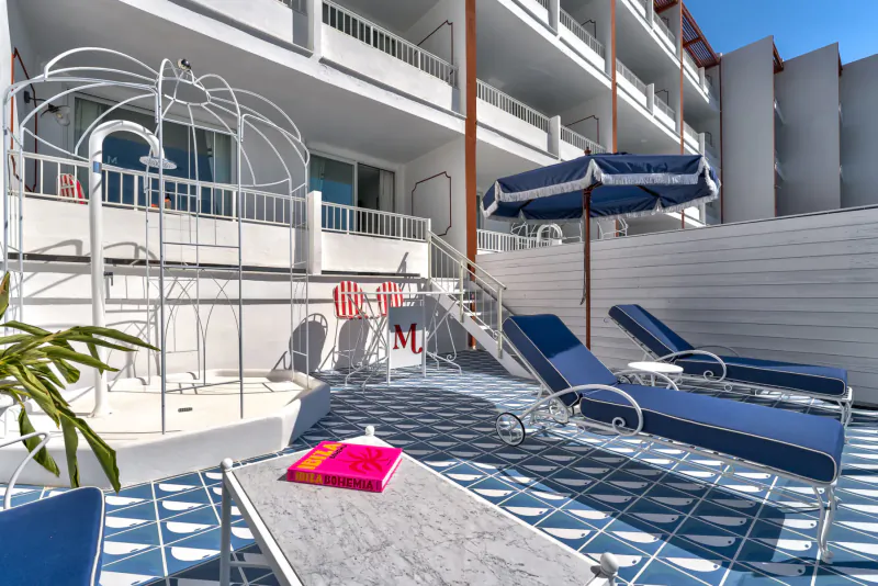 Luxury poolside terrace at Mongibello Hotel, Ibiza: blue loungers, striped umbrella, pink book on table, white hotel building.