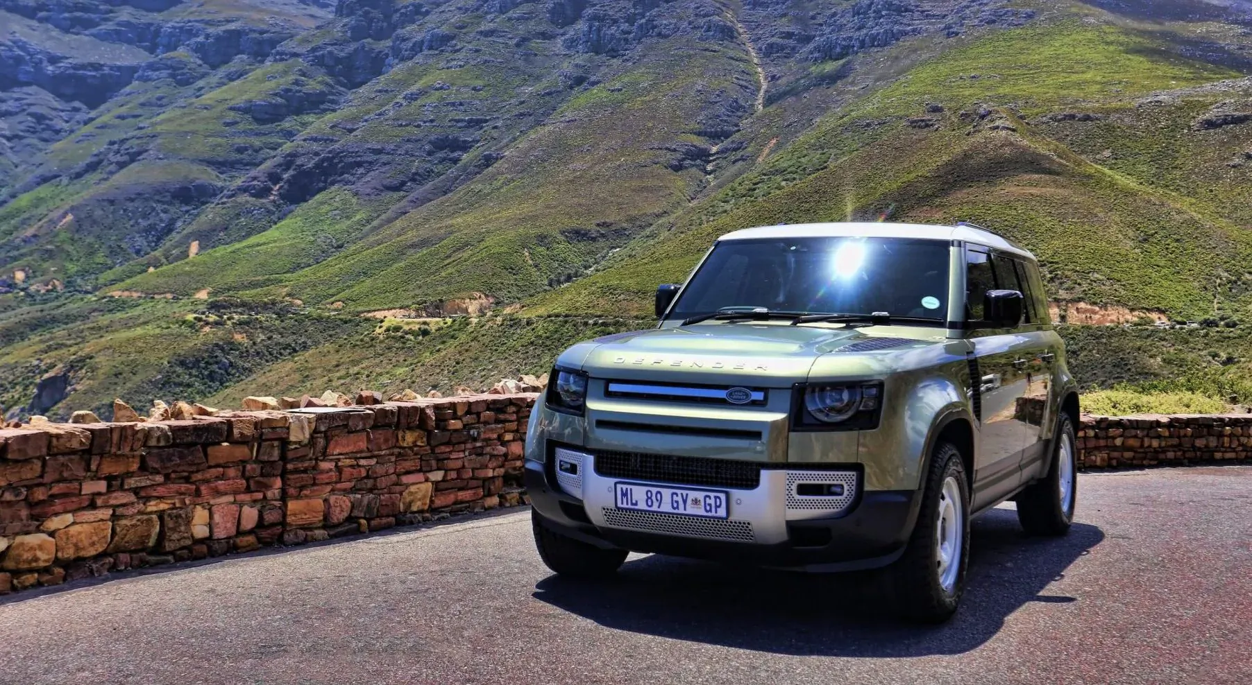 Green 2025 Land Rover Defender 110 parked on mountain road overlook with green hills and stone wall.