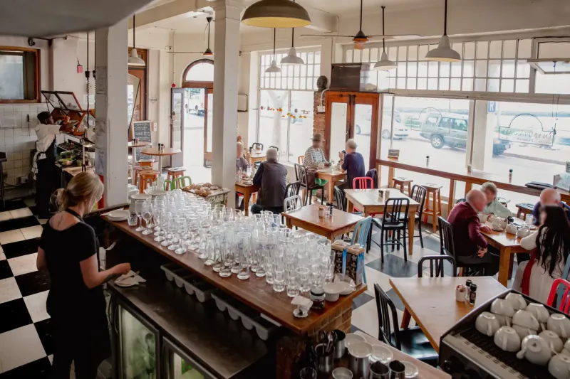 Busy Olympia Café in Kalk Bay: bartender behind wooden bar with glassware, patrons dining at tables, large windows overlooking street.