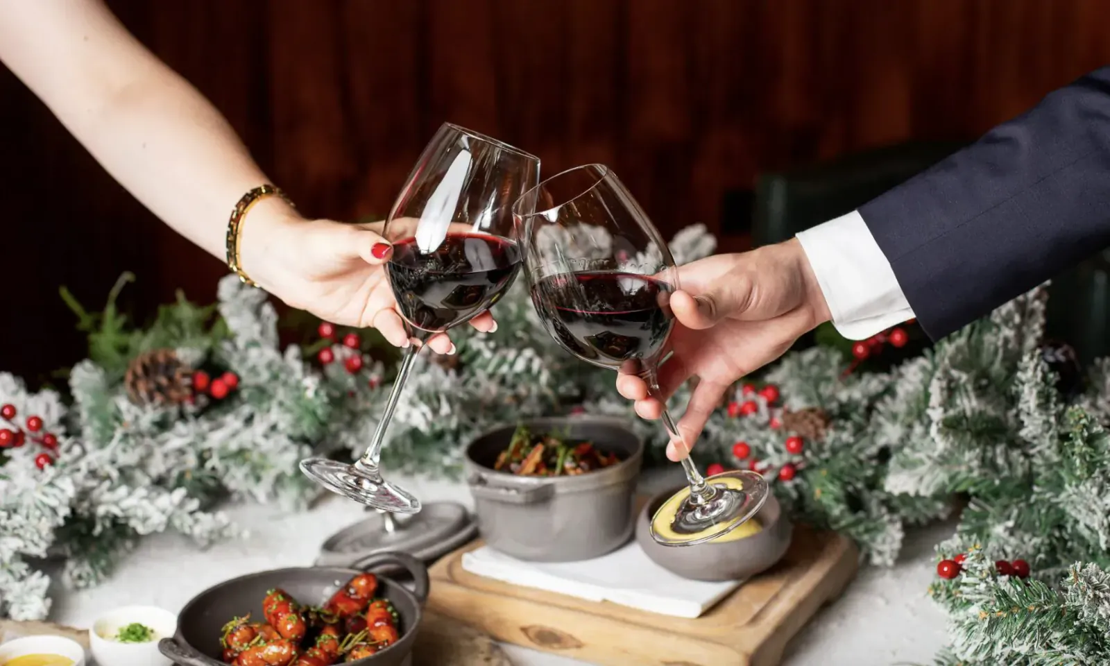 Couple toasting with red wine glasses amid Christmas greenery, beside gourmet dishes on wooden board