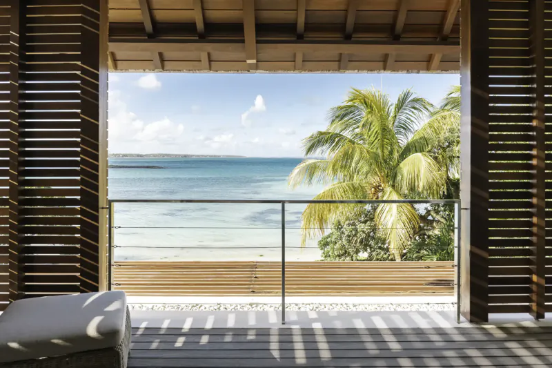 Open wooden shutters frame tropical beach view with turquoise sea, palm trees, and villa deck at Villas de Maitre Mauritius.