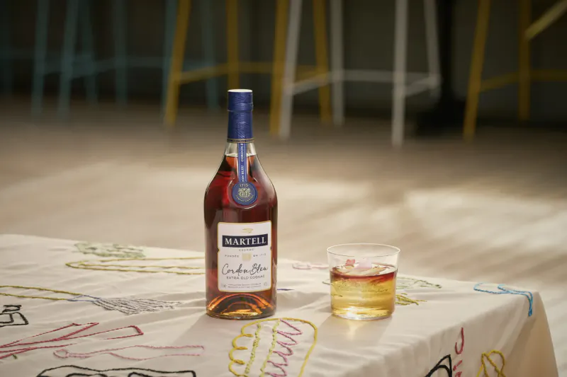Martell Cordon Bleu cognac bottle and glass on colorful table at L’Indigo bar, yellow stools in background.
