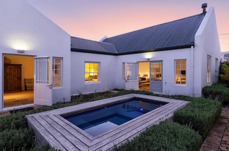 White Vishuis cottage with lit windows, blue plunge pool, and garden at sunset.