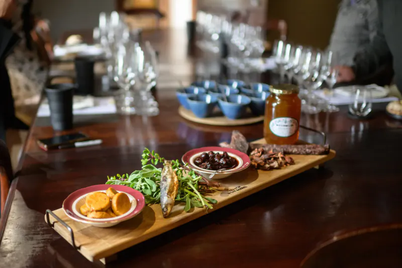 Wooden charcuterie board with orange slices, olives, anchovies, greens, honey jar, and blue bowls on a winery dining table