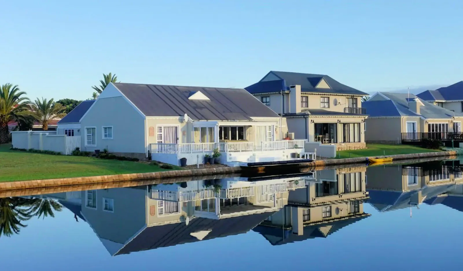 Row of modern white houses with blue roofs reflected in calm canal, palm trees and green lawns, sunny day.