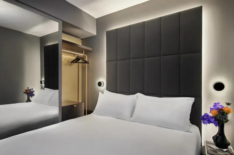 Modern hotel bedroom with gray upholstered headboard, white bed, wardrobe mirror, and purple flower vases on nightstands.