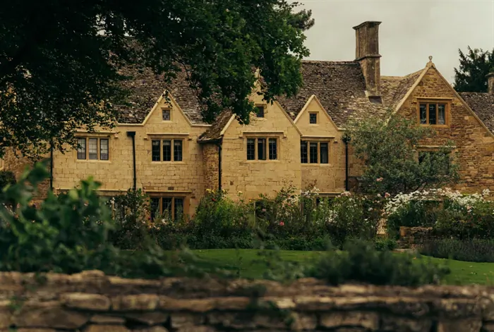 Stone Cotswold manor house with gabled roof, tall trees, rose garden, and stone wall in front, overcast sky.