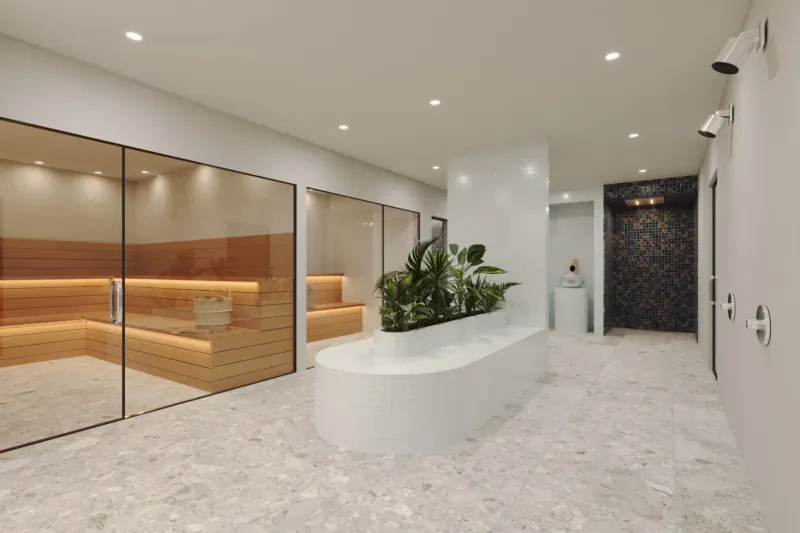 Modern luxury hotel spa hallway with glass-walled wooden saunas, potted plants, and curved white bench.