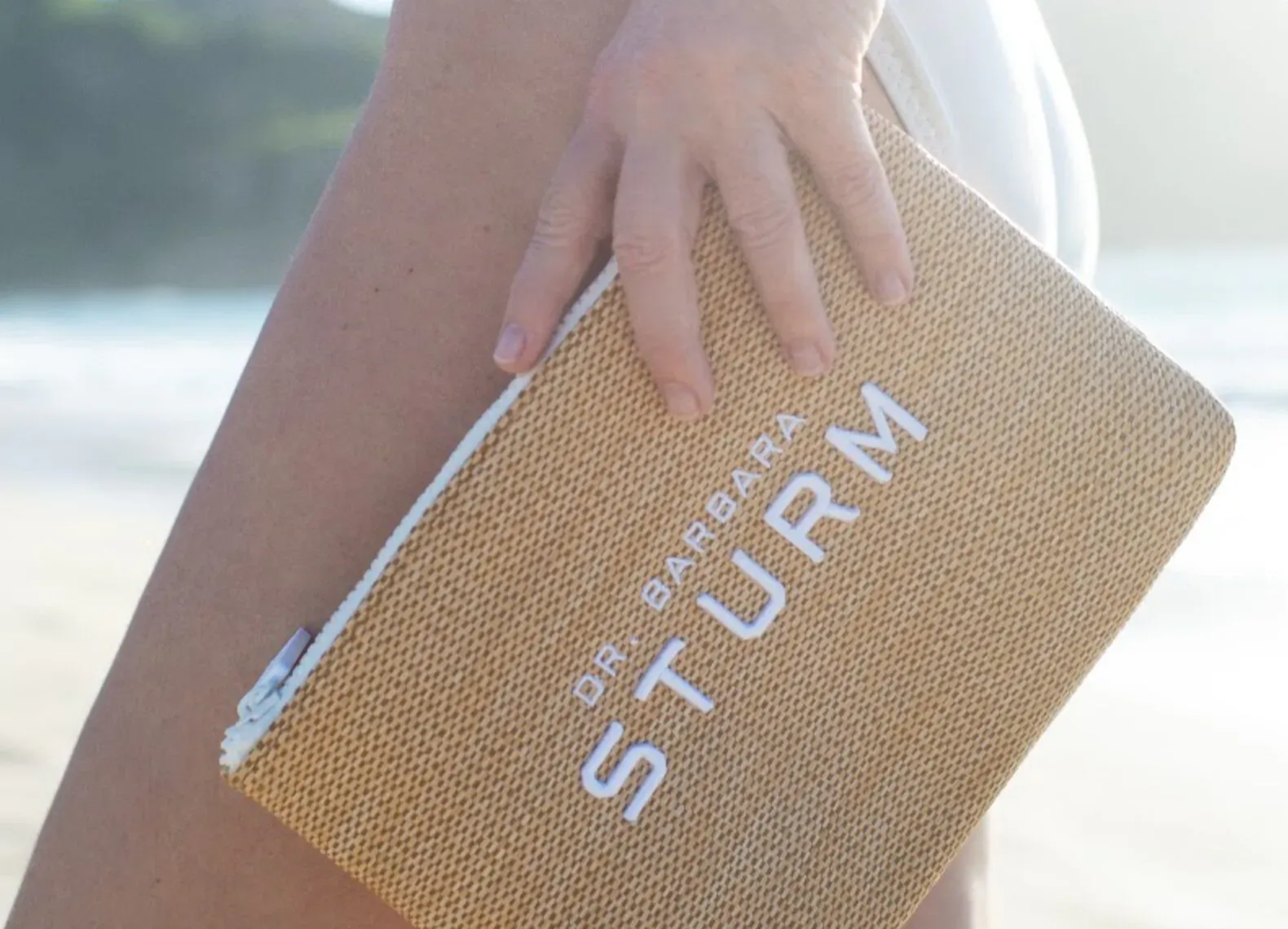 Woman in white swimsuit holding tan woven 'Dr. Sturm' pouch on sunny beach